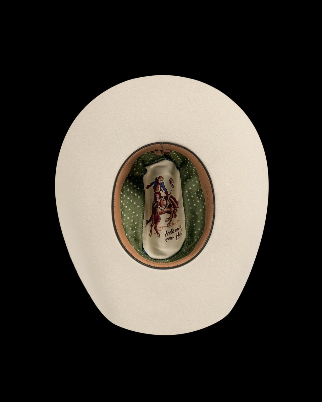 Top view of a beige cowboy hat with a green polka dot lining and an illustration of a cowboy riding a bucking horse on the inside crown.