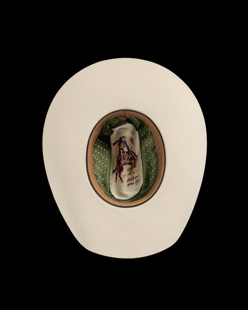 Top view of a beige cowboy hat with a green polka dot lining and an illustration of a cowboy riding a bucking horse on the inside crown.