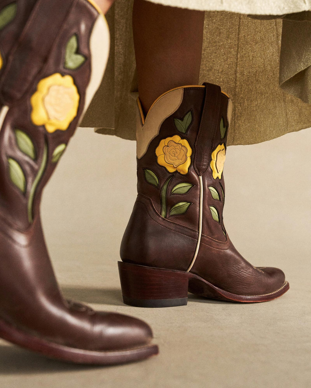The Jolene in Truffle Cowhide. Dark brown truffle cowhide cowboy boots with yellow rose and green leaf appliqué, worn with an olive skirt. A taller matching boot is visible in the foreground.