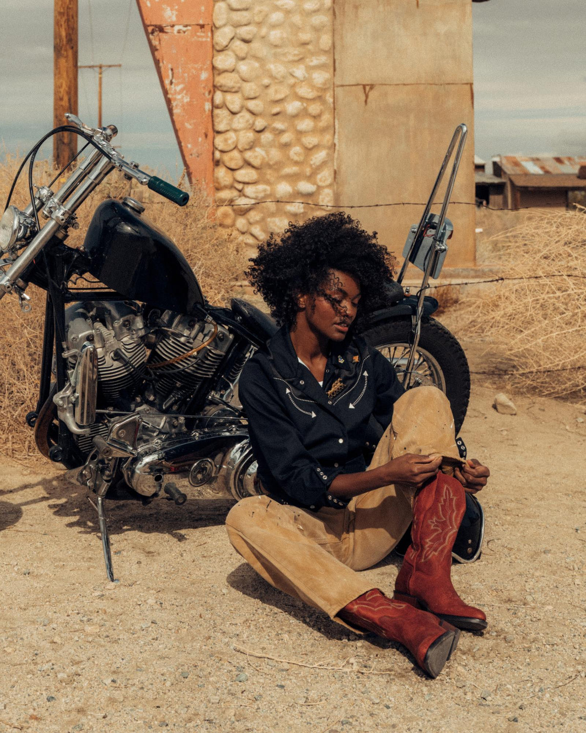 The Britt in Redwood Goat. A person wearing a black shirt, tan pants, and red boots sits on the ground next to a motorcycle in a rustic, desert-like setting, channeling the adventurous spirit of the Redwood Goat.