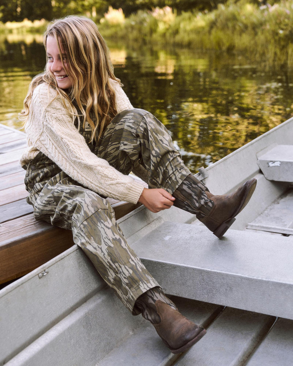 A person with long hair in a sweater and camouflage pants sits on a dock by the water, smiling and adjusting their brown boots while sitting on the edge of a metal boat.