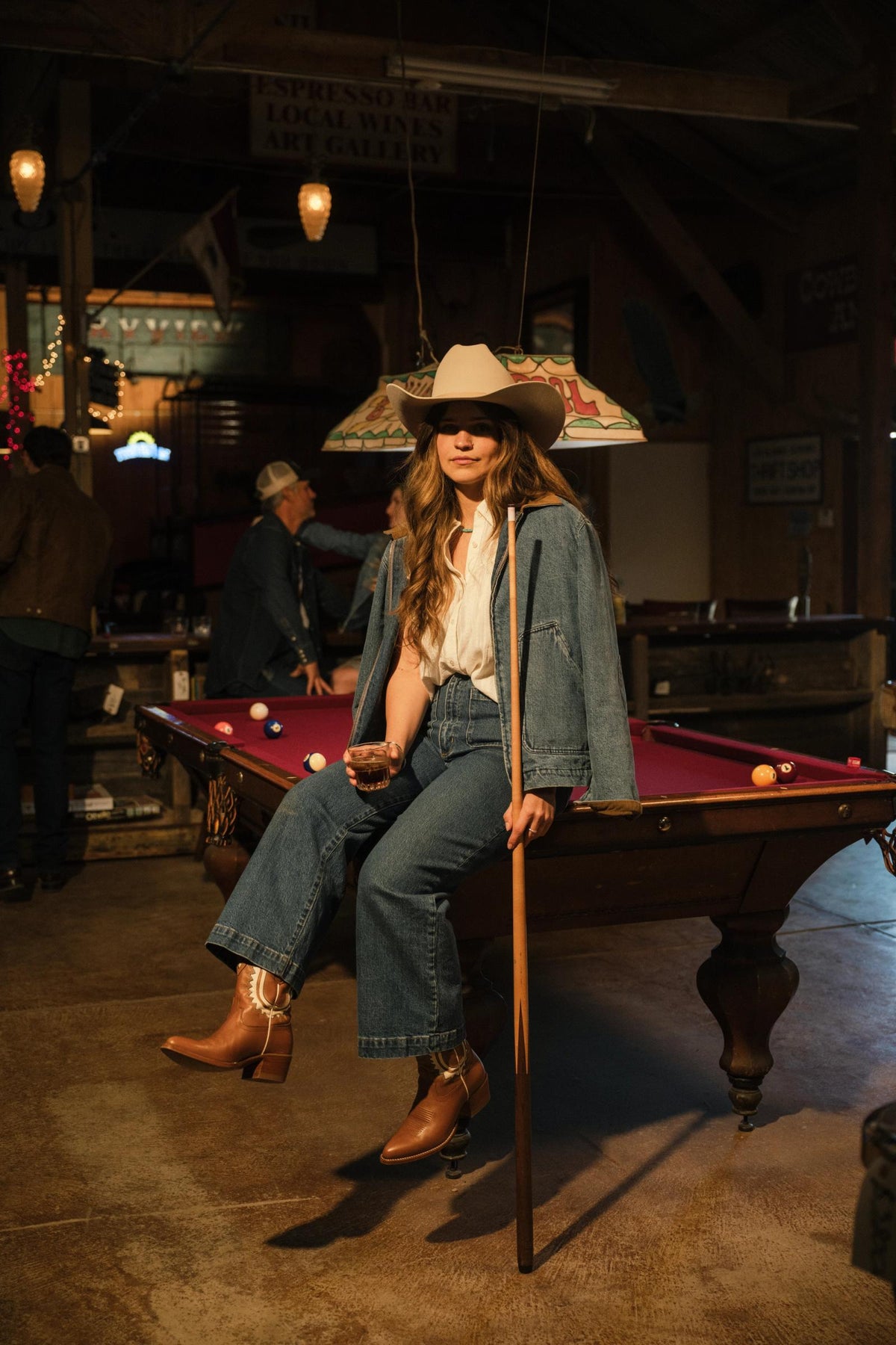 A person in a cowboy hat and denim outfit sits on a pool table holding a cue stick in a dimly lit room.