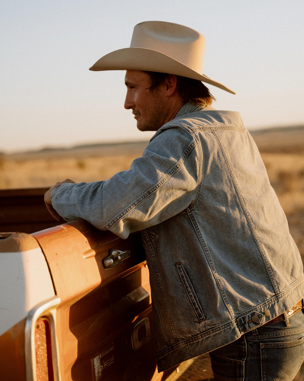 A man in a denim jacket and cowboy hat leans on the back of a dusty pickup truck in a dry, open landscape.