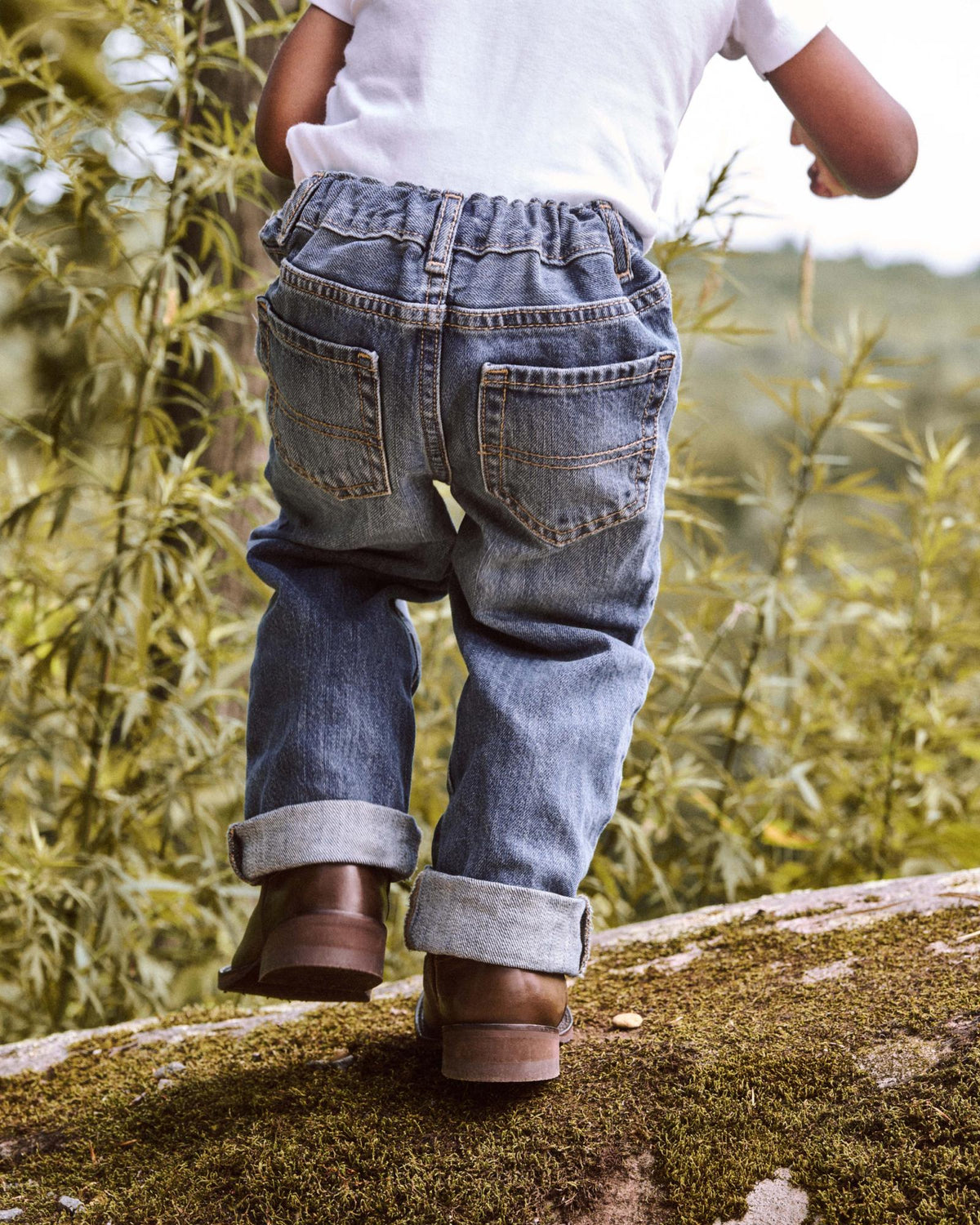 A child in a white shirt, rolled-up jeans, and brown boots stands on a mossy rock outdoors with greenery in the background.