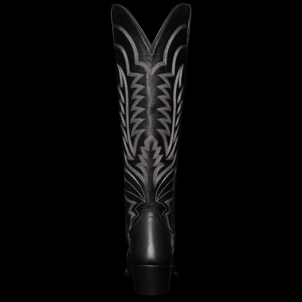 A tall, black cowboy boot with intricate white stitching and a pointed toe, viewed from the front.