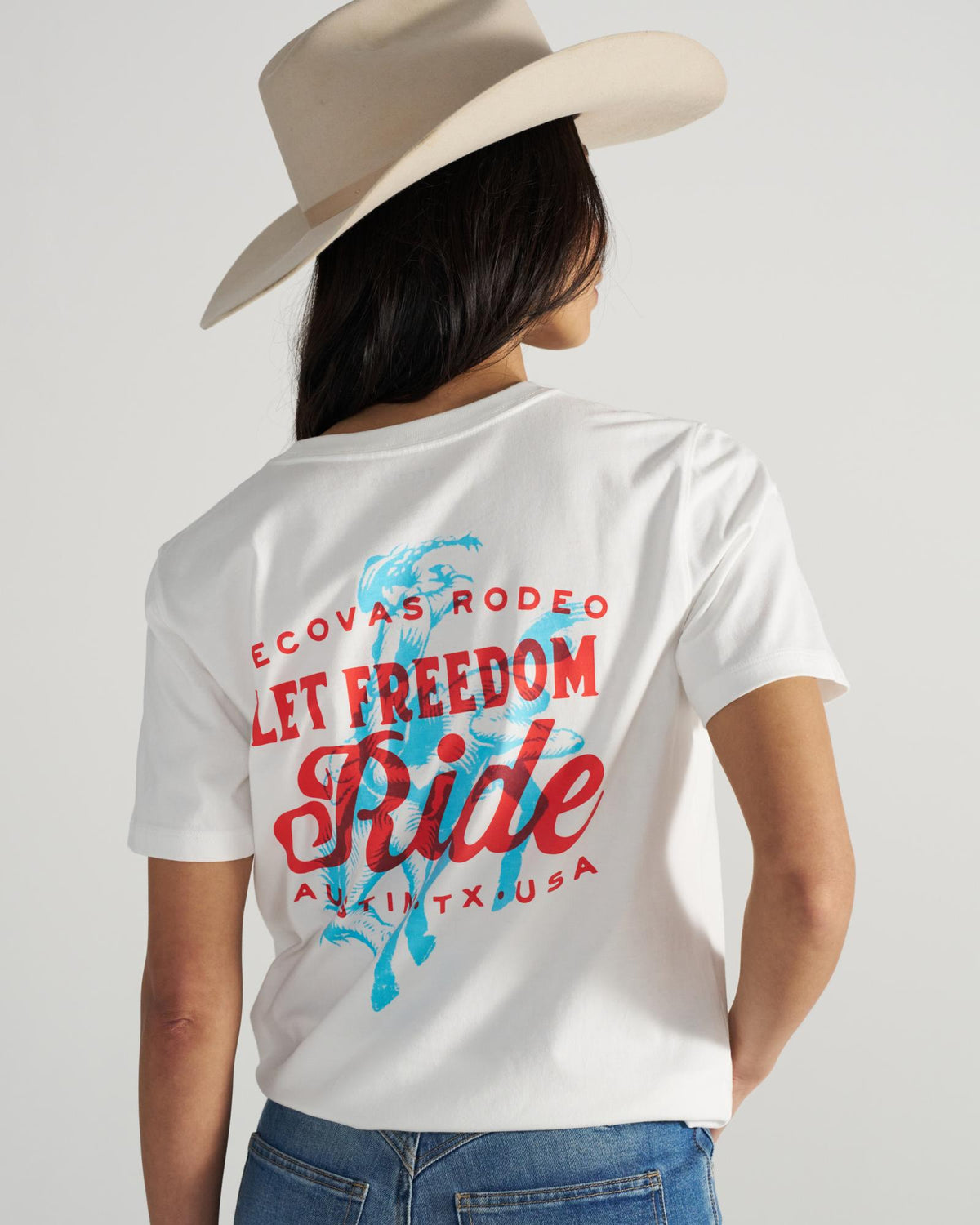 Person wearing a white cowboy hat and a white T-shirt with red and blue text and graphics that says "Let Freedom Ride" and "ECOVAS RODEO AUSTIN TX USA" on the back.