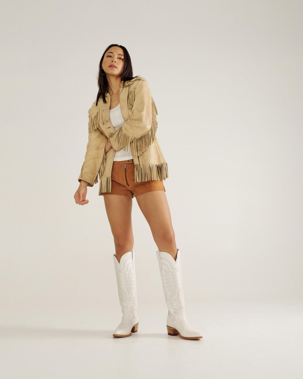 A woman stands against a plain background wearing a fringe jacket, white top, brown shorts, and white cowboy boots.