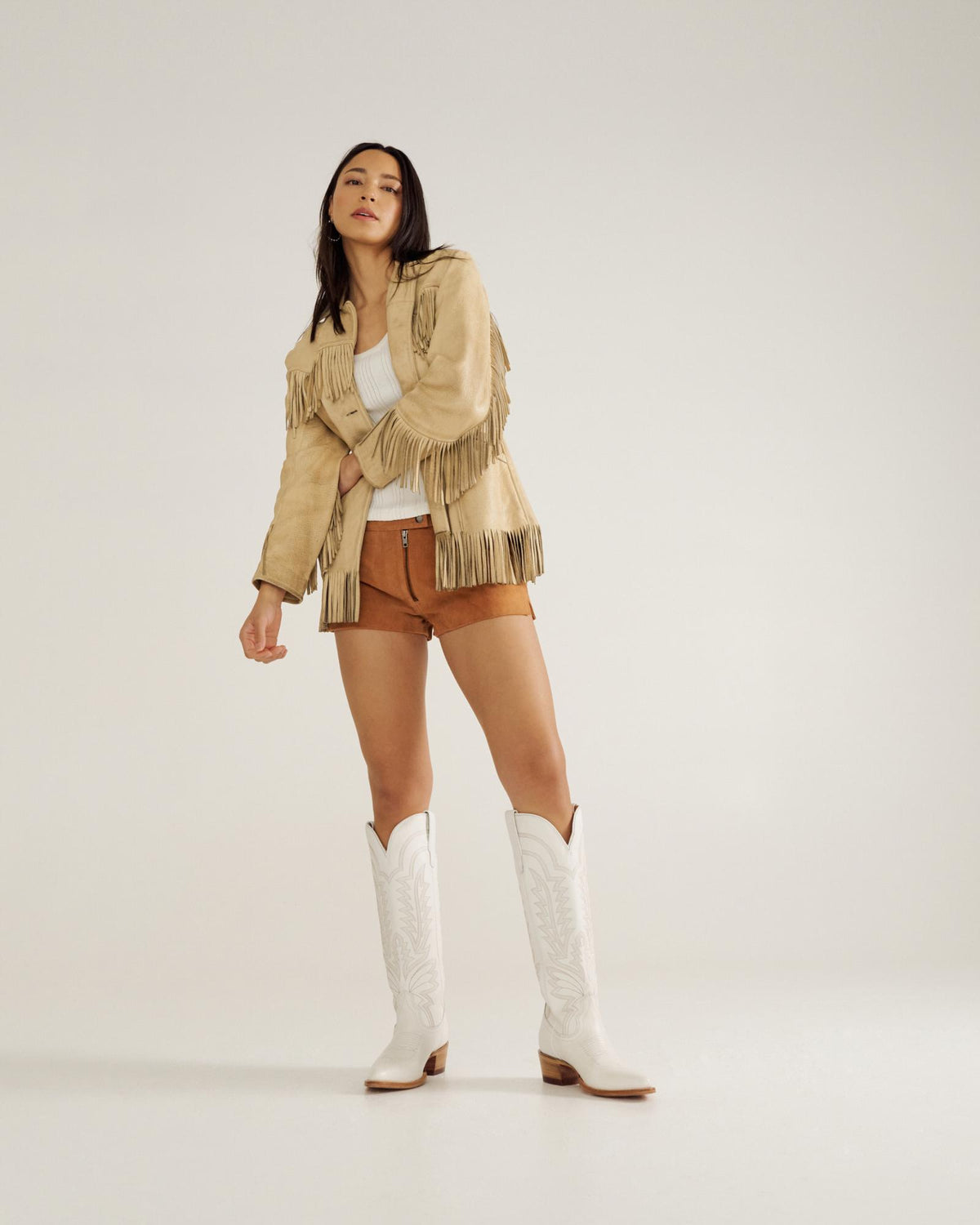 A woman stands against a plain background wearing a fringe jacket, white top, brown shorts, and white cowboy boots.