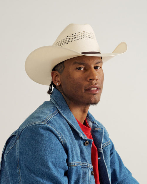 A man wearing a white cowboy hat, denim jacket, and red shirt looks toward the camera against a plain background.