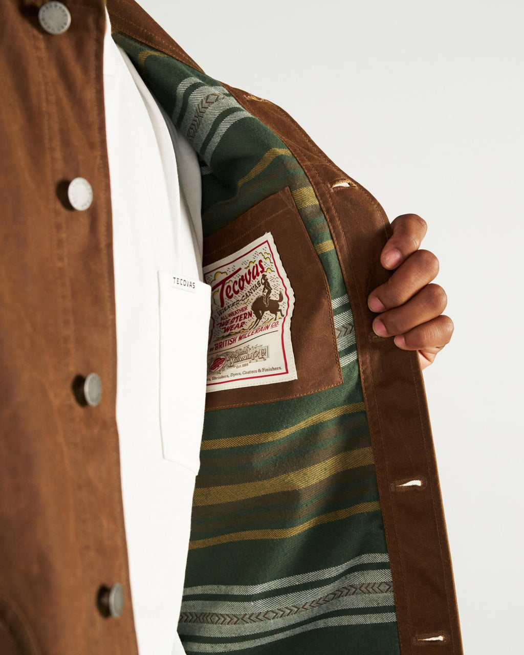 A person wearing a brown jacket with a green striped interior lining and a Tecovas brand patch sewn into the chest pocket area.