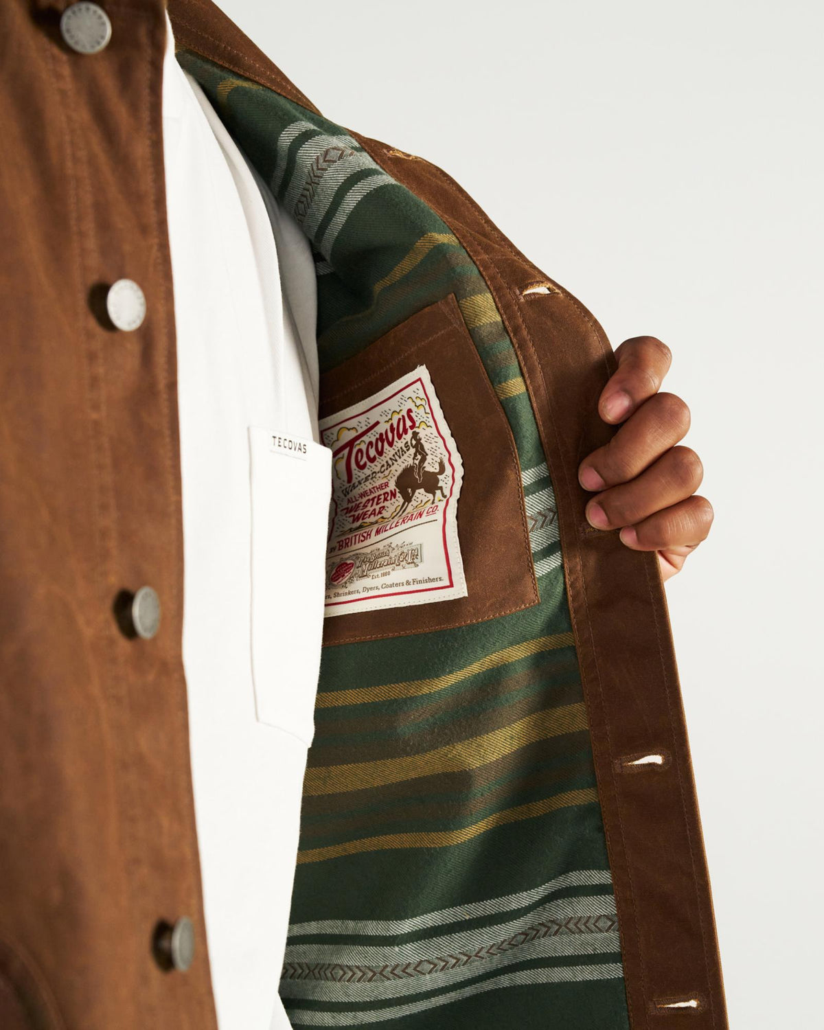 A person wearing a brown jacket with a green striped interior lining and a Tecovas brand patch sewn into the chest pocket area.