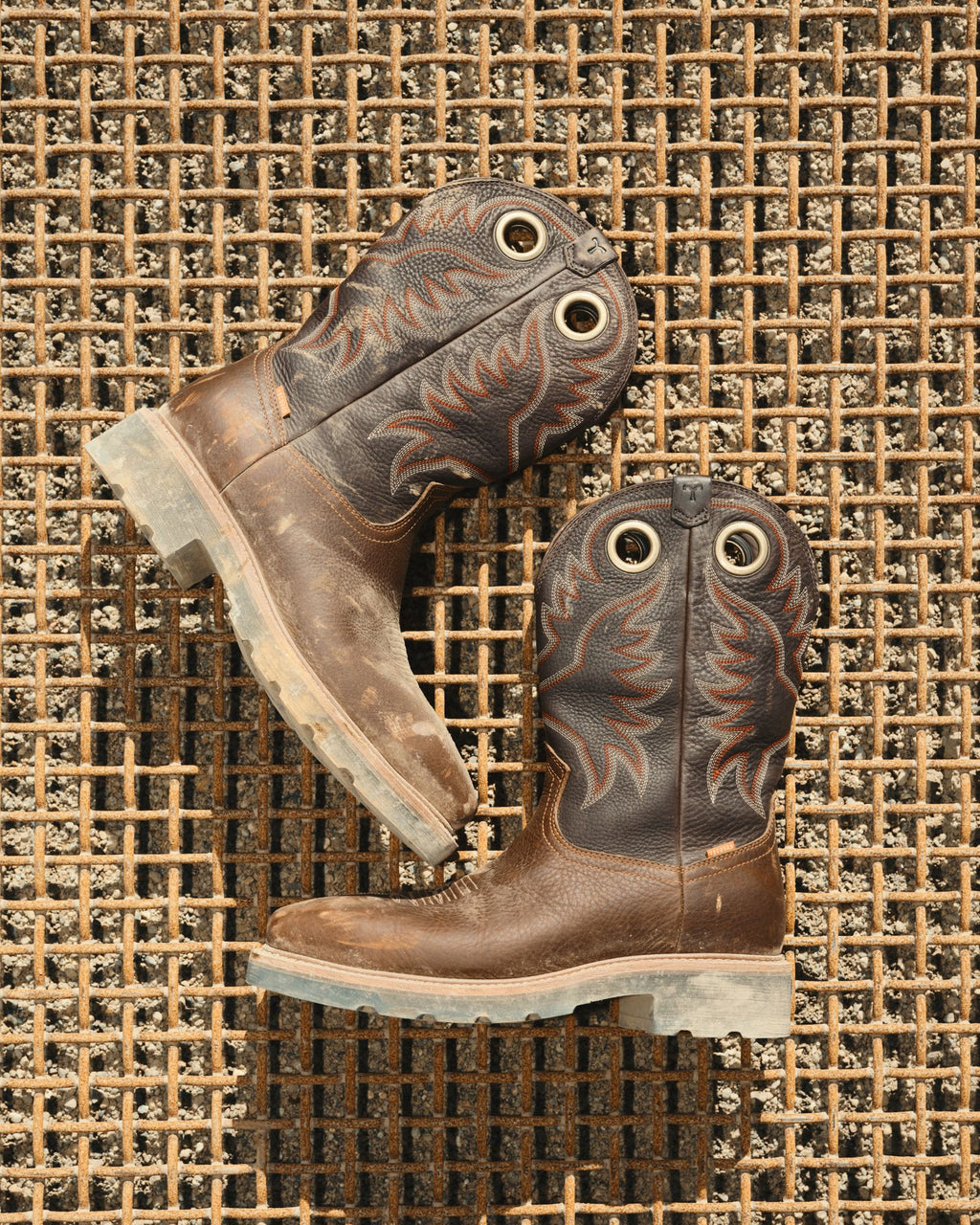 LongHaul Waterproof Square Composite Toe Work Boot in Saddle/Fossil Cowhide. A pair of worn maple cowhide cowboy boots with black and orange stitching rests on a rusty metal grid surface.