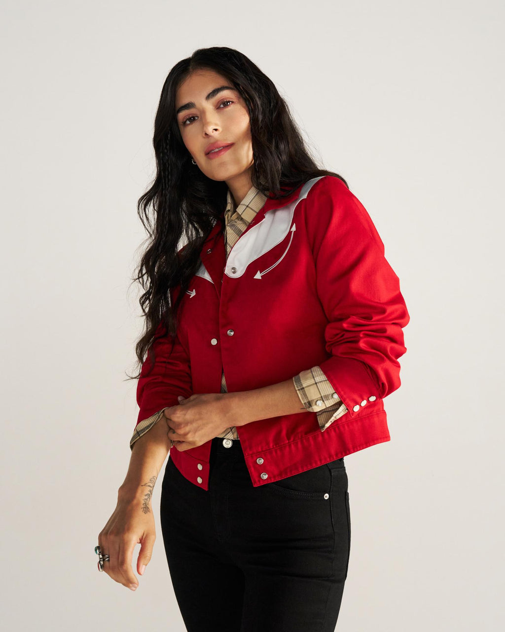 A woman with long dark hair wears a red jacket over a plaid shirt and black pants, standing against a plain light background.
