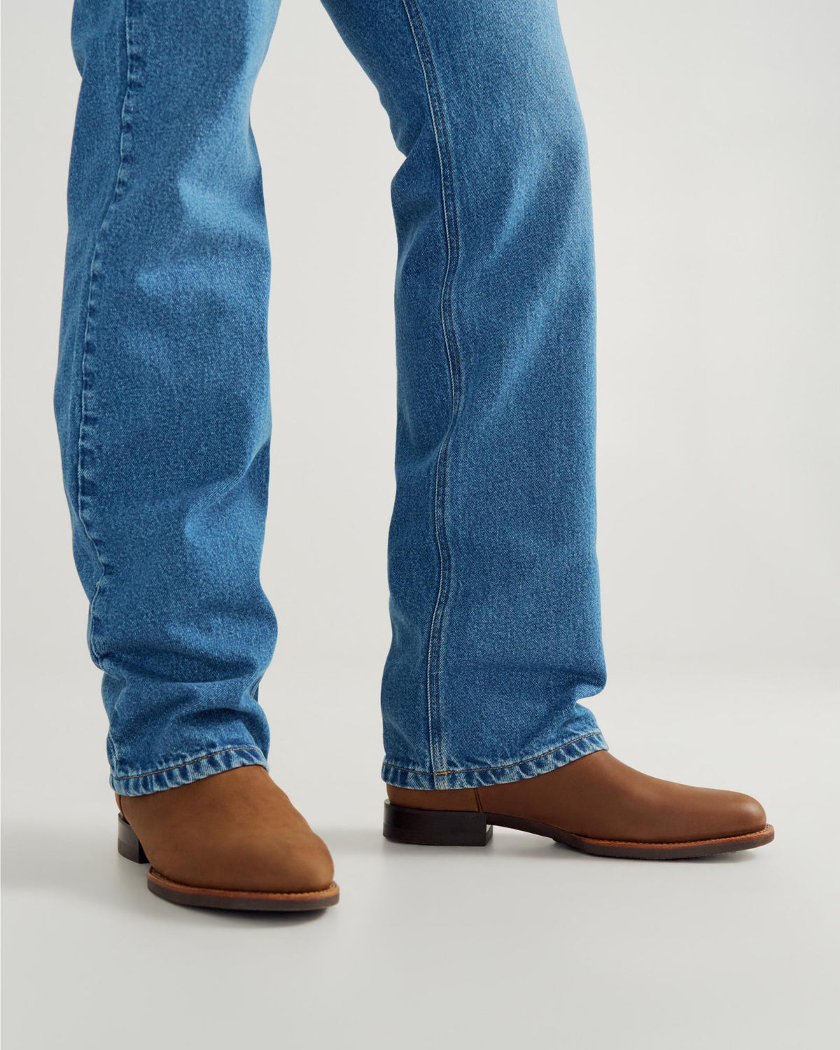 The Glen in Khaki Cowhide. Person wearing blue jeans and brown leather boots, standing on a light-colored surface, showcases classic style with a hint of cowhide charm.