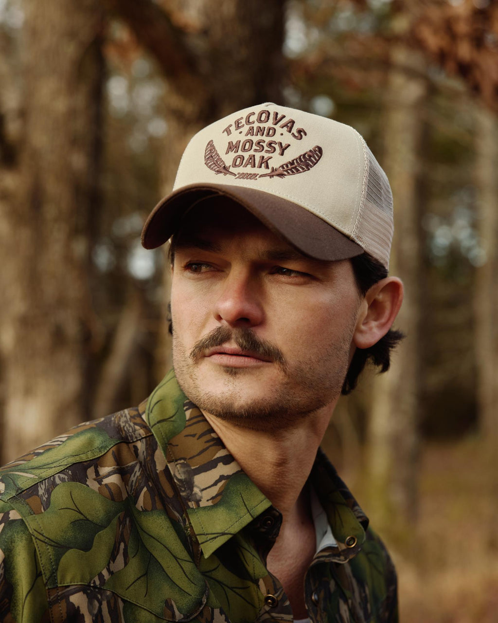 A man with a mustache wearing a camouflage shirt and a beige cap that reads "Tecovas and Mossy Oak" stands outdoors in a wooded area.