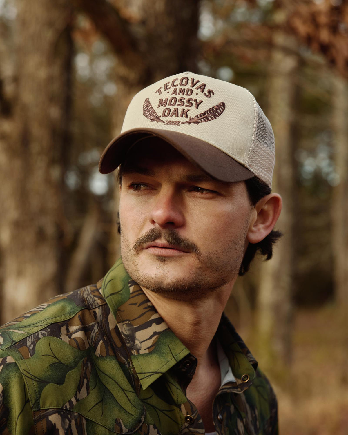 A man with a mustache wearing a camouflage shirt and a beige cap that reads "Tecovas and Mossy Oak" stands outdoors in a wooded area.