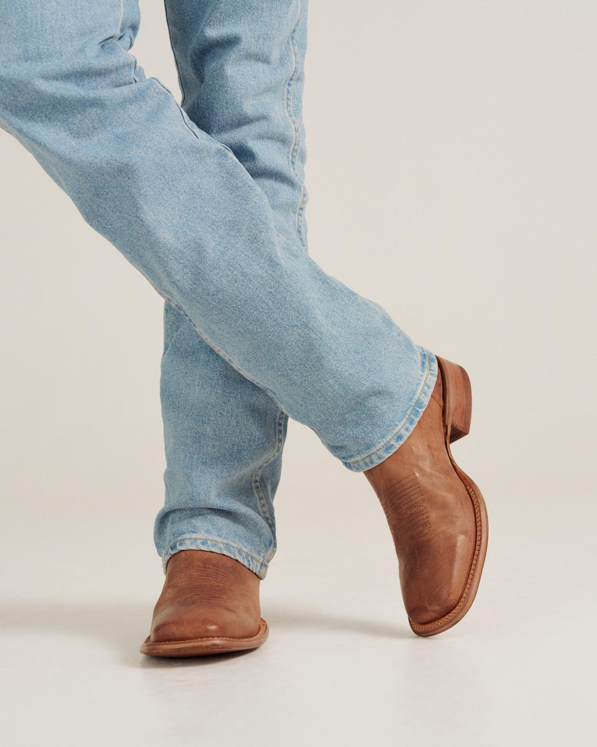 The Doc in Scotch Goat. Person wearing light blue jeans and brown leather boots from The Doc Goat stands with legs crossed on a plain background.