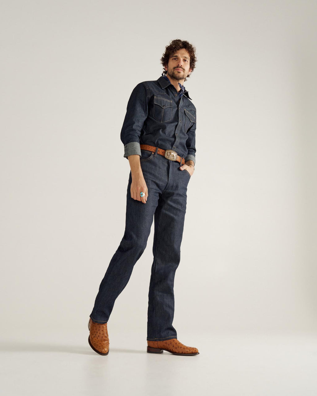 A man stands against a plain background wearing a dark denim shirt and jeans, a brown belt, brown cowboy boots, and a turquoise ring.