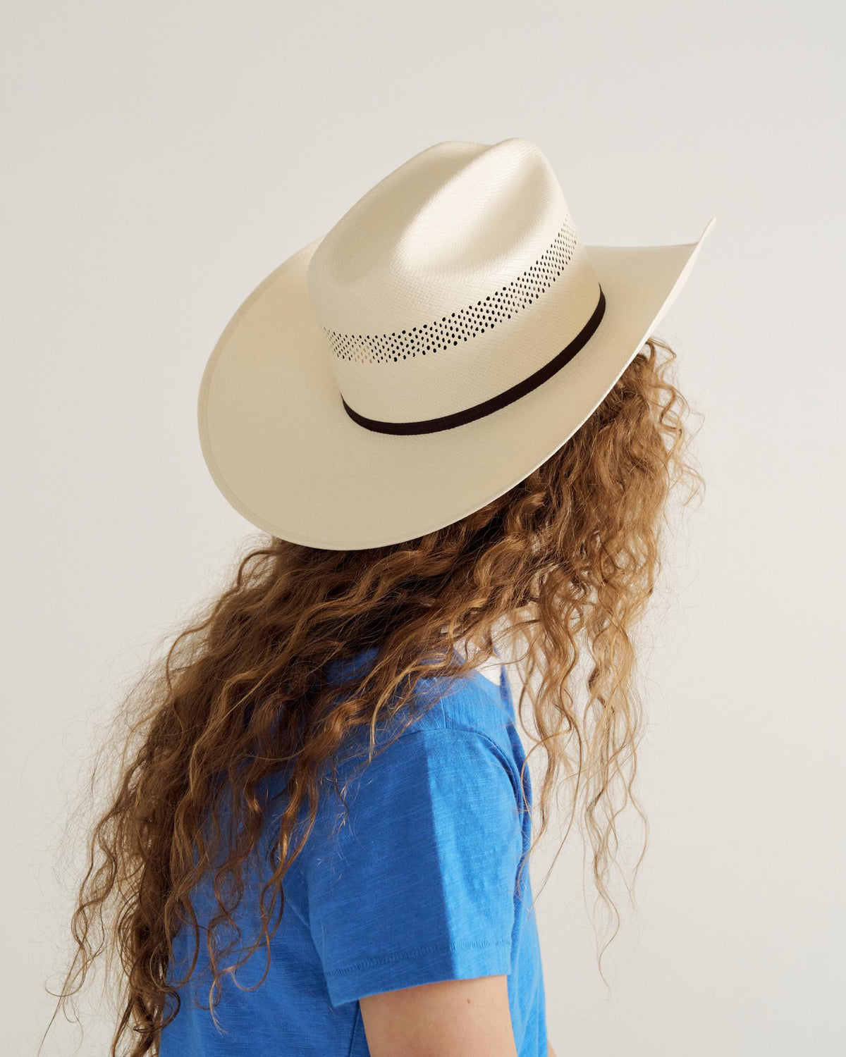 Person with long, curly hair wearing a light-colored cowboy hat and a blue T-shirt, facing away from the camera against a plain background.