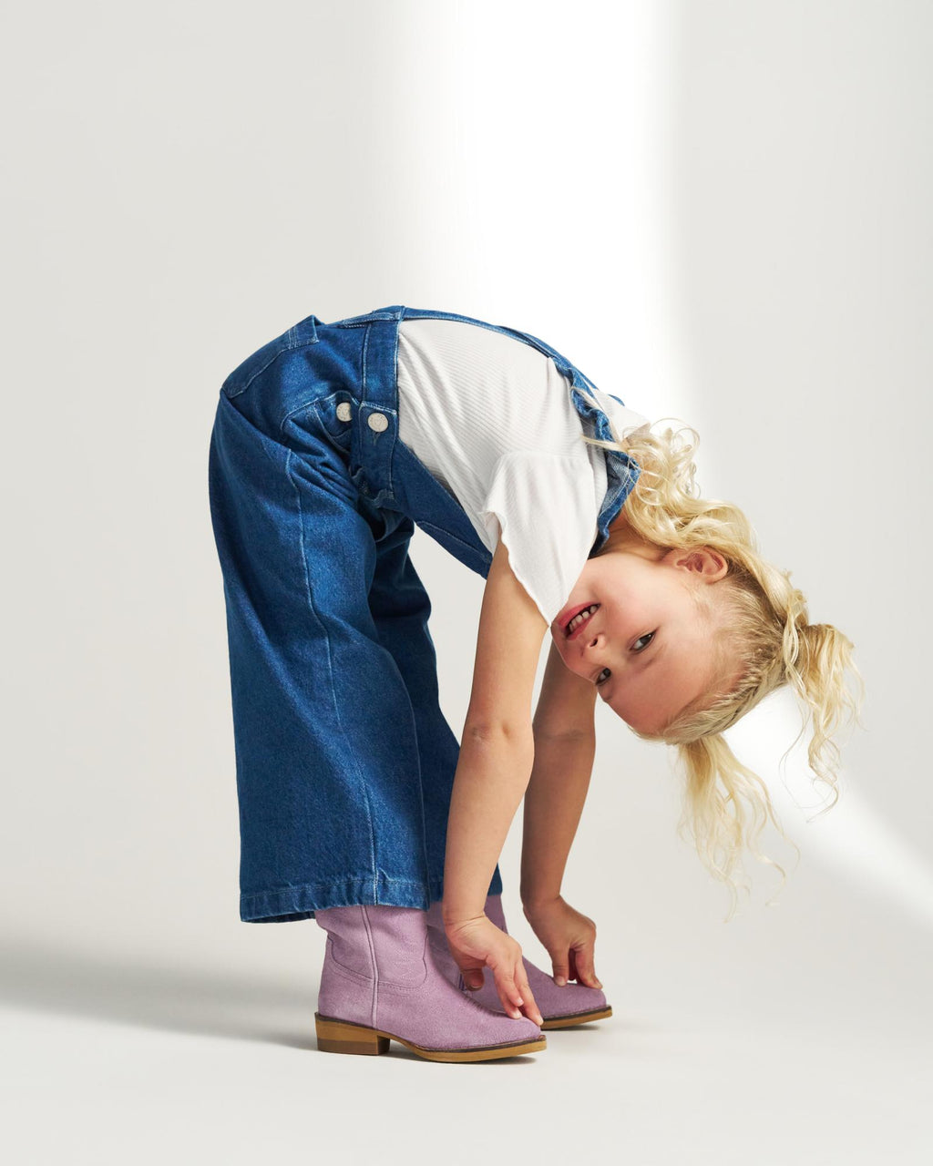 A young child with curly blonde hair bends over, touching lavender boots, wearing denim overalls and a white t-shirt against a plain background.
