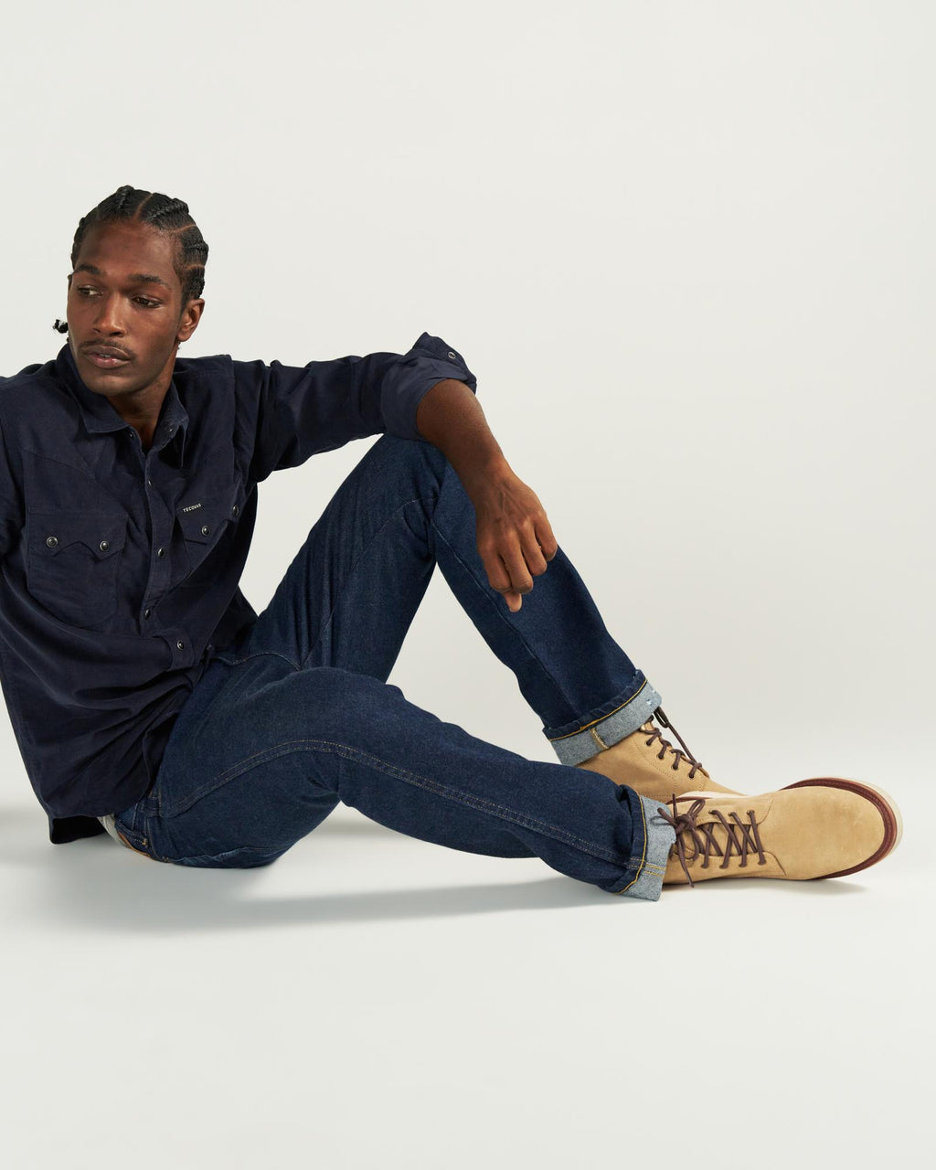 The Knox Lace Up Shearling in Sandstone Roughout. Man sitting on floor with one knee up, wearing a dark button-up shirt, rolled-up jeans, and tan Sandstone Roughout lace-up boots, against a plain light background.