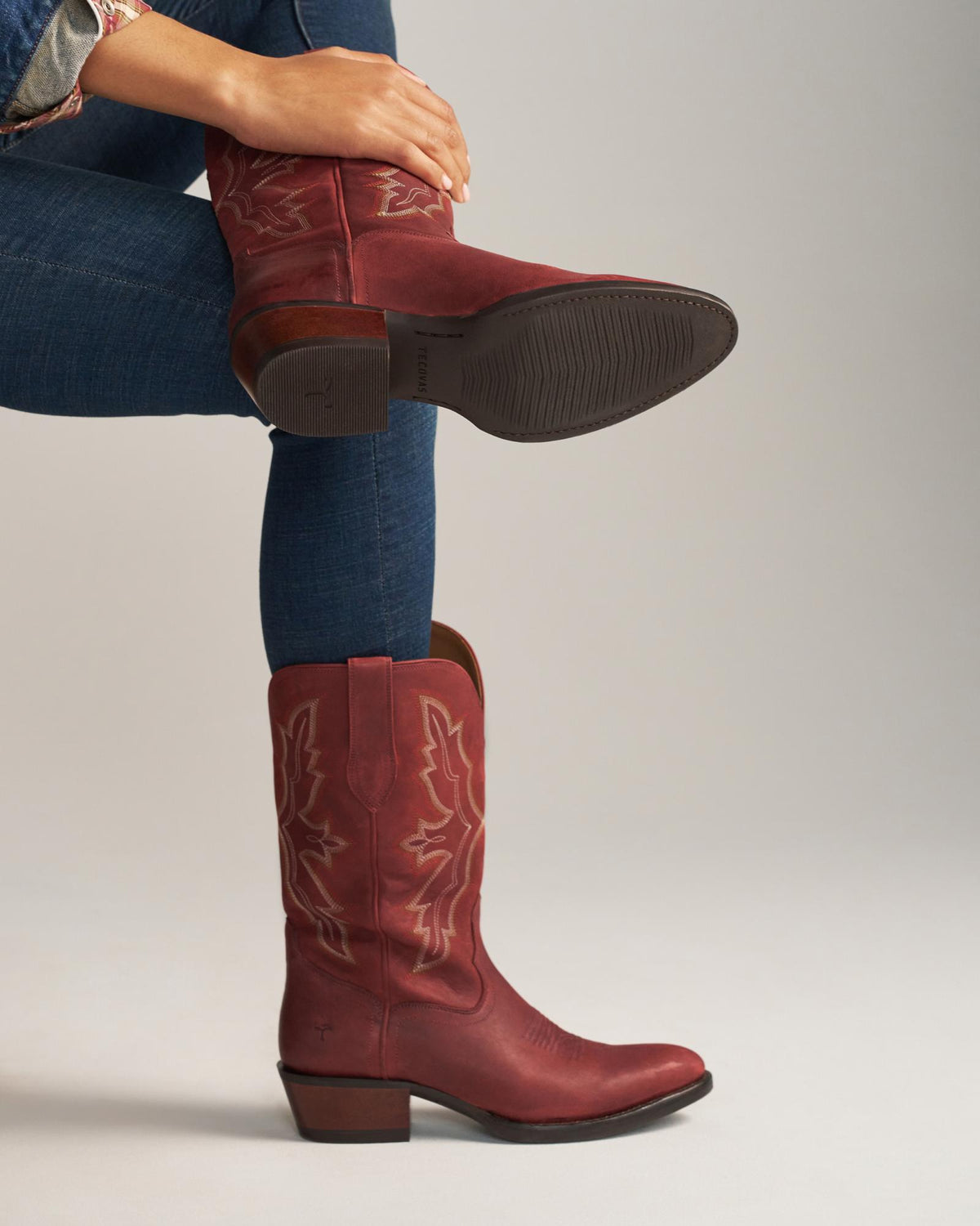 The Britt in Redwood Goat. A person wearing blue jeans and a patterned shirt models a pair of Britt Goat red cowboy boots with decorative stitching, crossing one leg over the other.