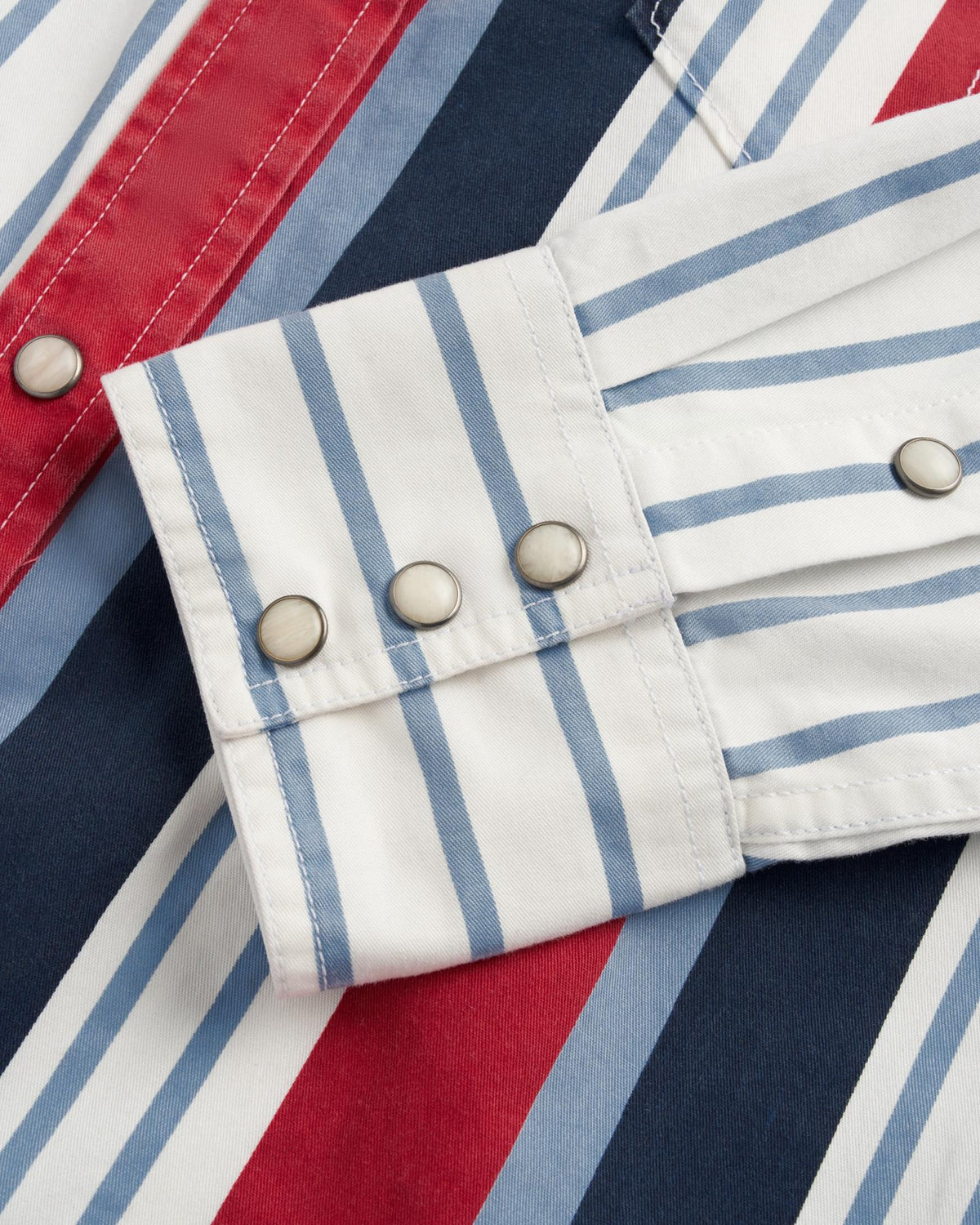 A close-up of a striped shirt cuff with three silver snap buttons. The shirt features vertical blue, white, red, and navy stripes.