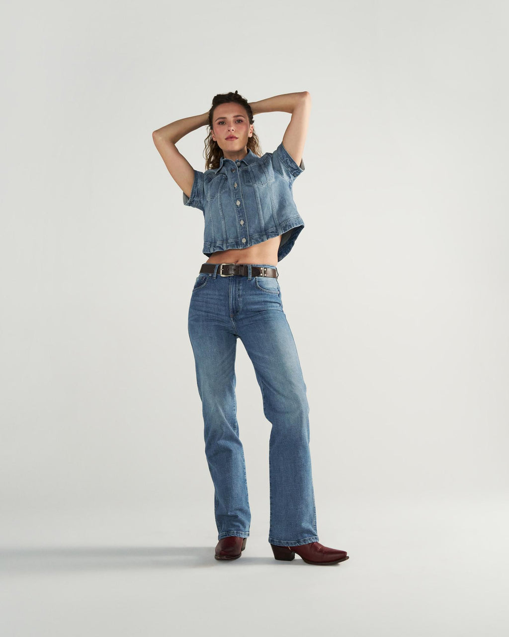 A person stands against a plain background wearing a short-sleeve denim shirt, blue jeans, a black belt, and brown boots, with arms raised and hands behind their head.