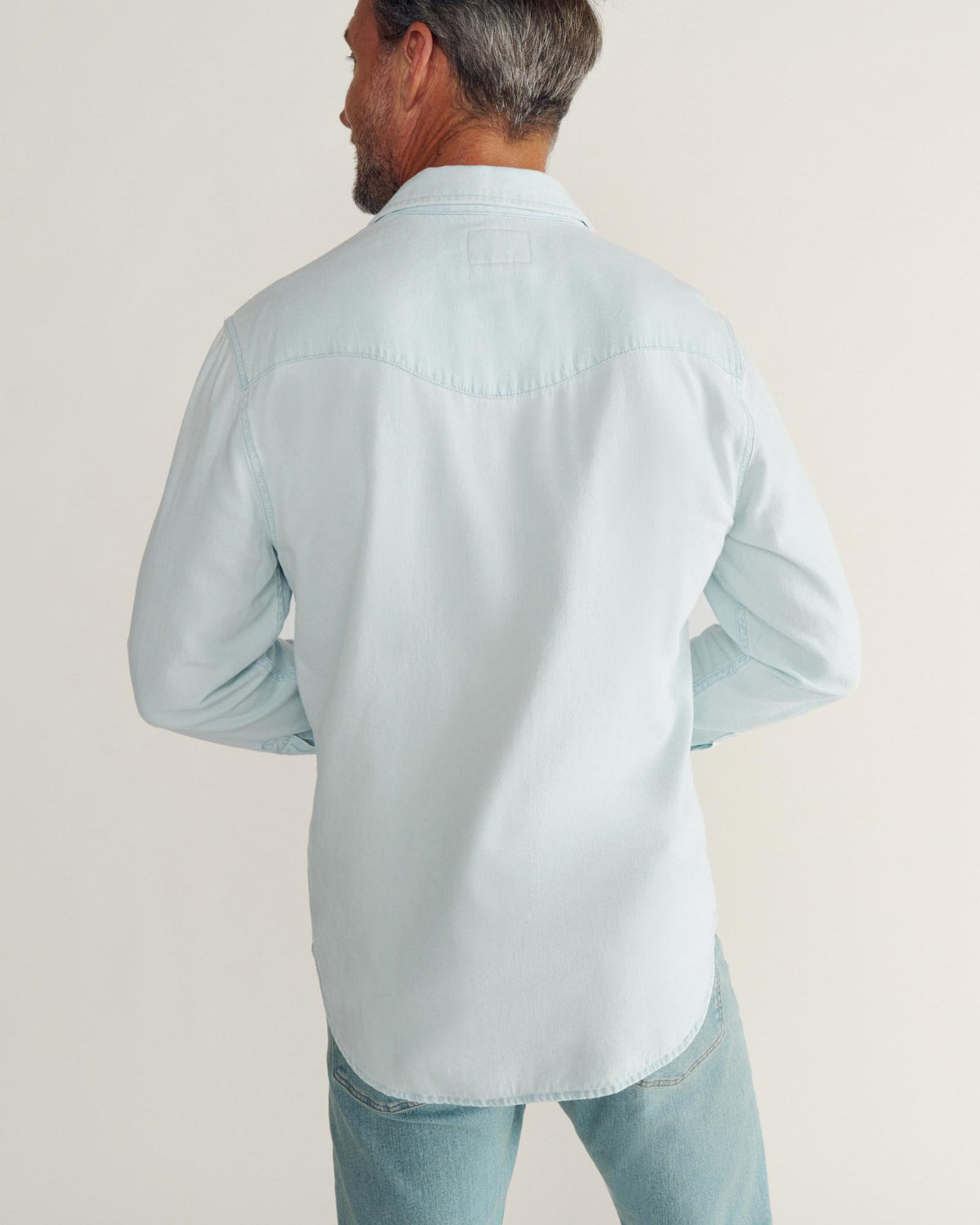 A man wearing a light blue denim shirt and jeans stands facing away from the camera against a plain background.