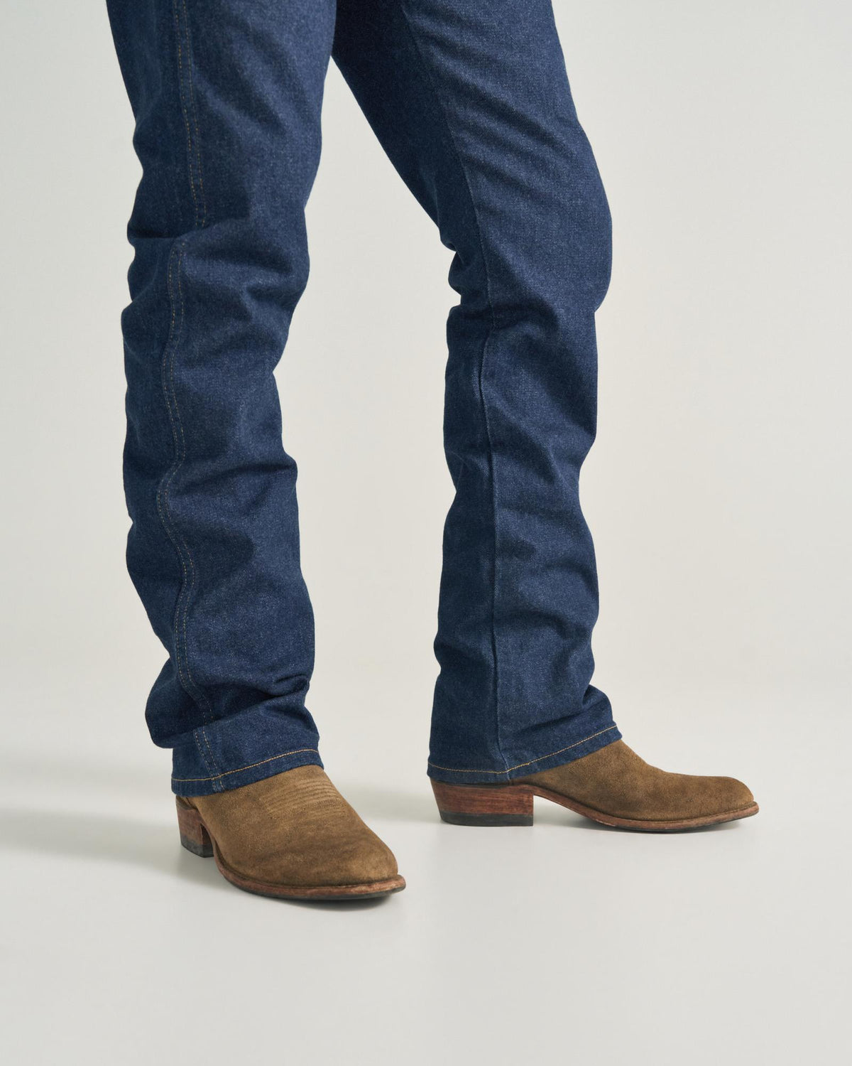 The Johnny in Gravel Suede. Wearing blue jeans and brown Gravel Suede cowboy boots, the person stands on a light-colored surface. Only their legs and feet are visible.