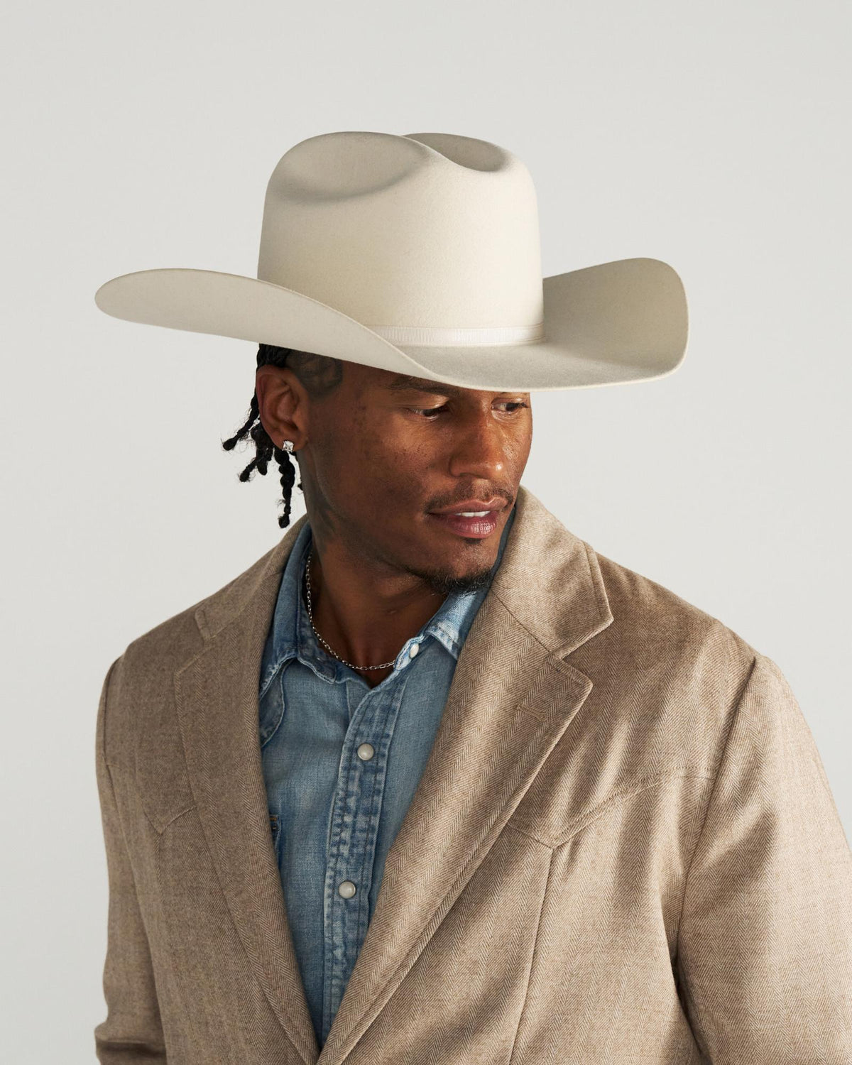 Man wearing a beige cowboy hat, tan suit jacket, and denim shirt, looking to the side against a plain light background.