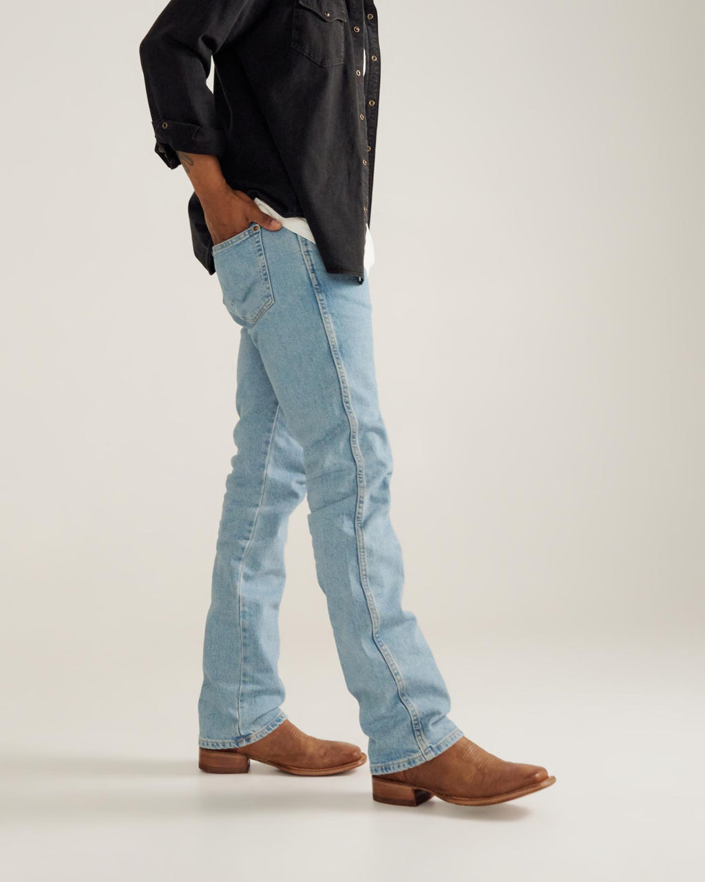 Person standing in light blue jeans, brown boots, and a dark long-sleeve shirt with hands in pockets against a plain background.