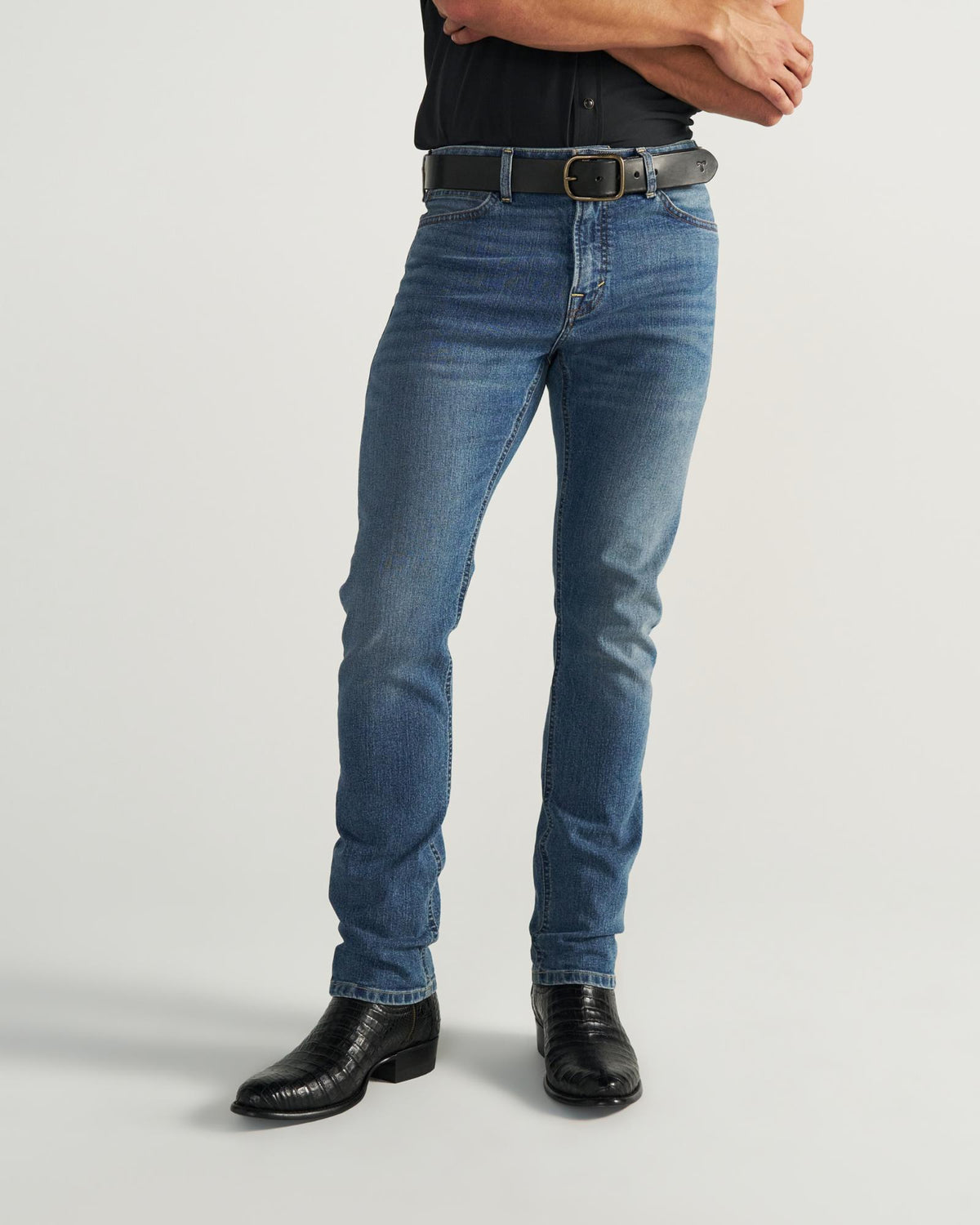 Person wearing blue jeans, a black belt, black shirt, and black cowboy boots stands with arms crossed against a plain background.