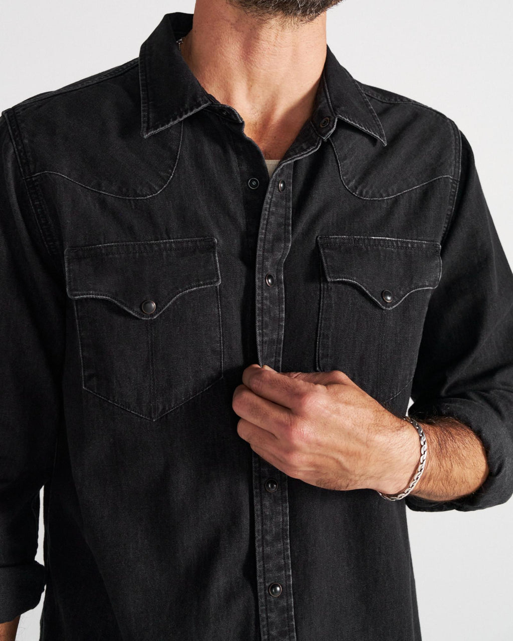 A man wearing a black denim button-up shirt with two chest pockets adjusts the front. Only his torso and part of his face are visible.