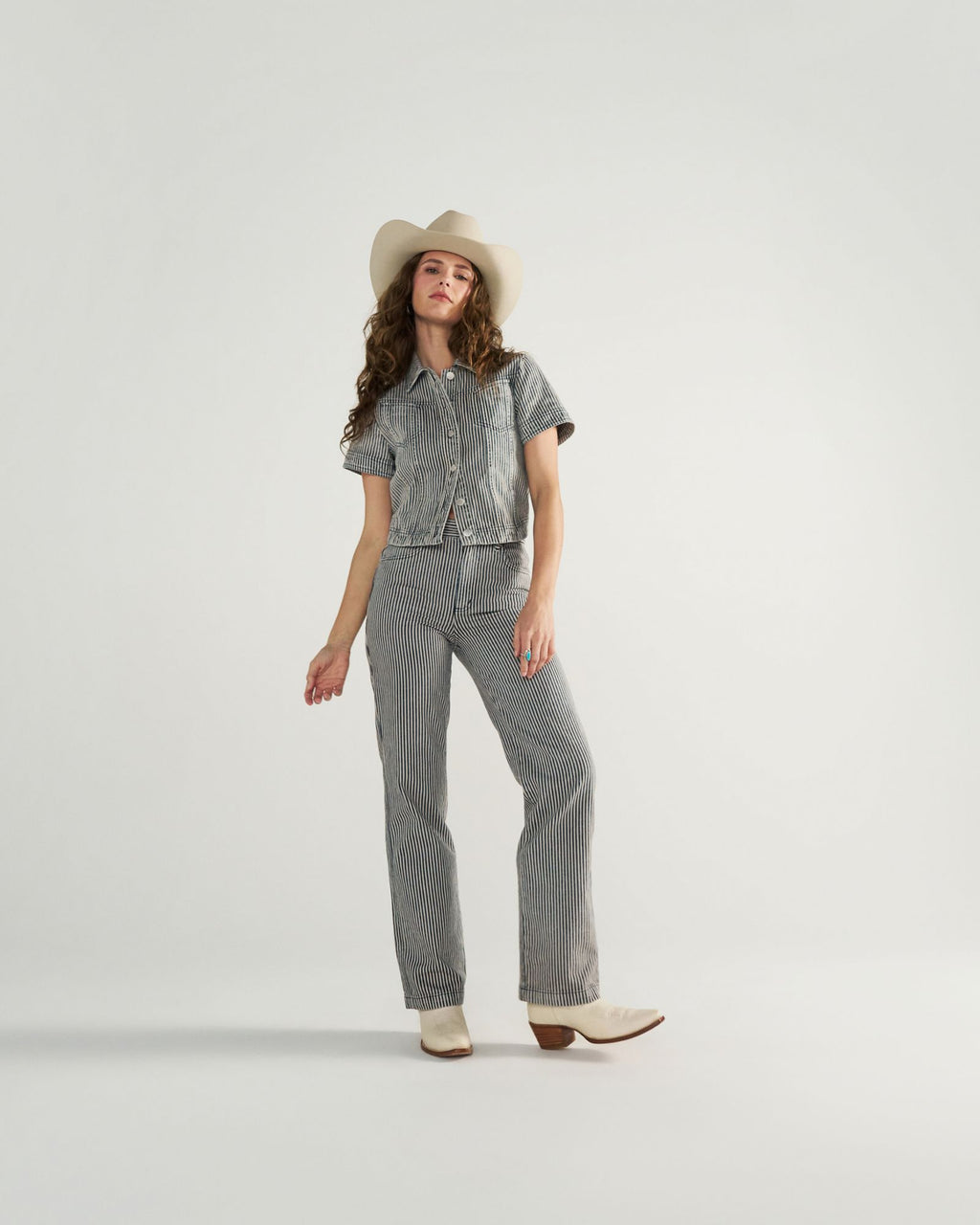 A woman wearing a white cowboy hat, short-sleeve striped button-up shirt, matching striped pants, and white boots stands against a plain white background.