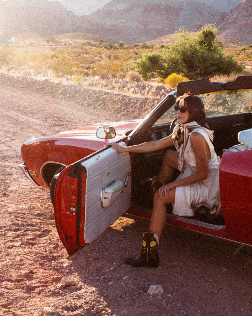 The Jolene in Truffle Cowhide. A person wearing a headscarf and sunglasses sits in a red convertible parked on a dirt road in a desert landscape, with the driver’s door open. Draped across the passenger seat is The Jolene Cowhide, its distinctive pattern catching the sunlight amidst the sandy backdrop.