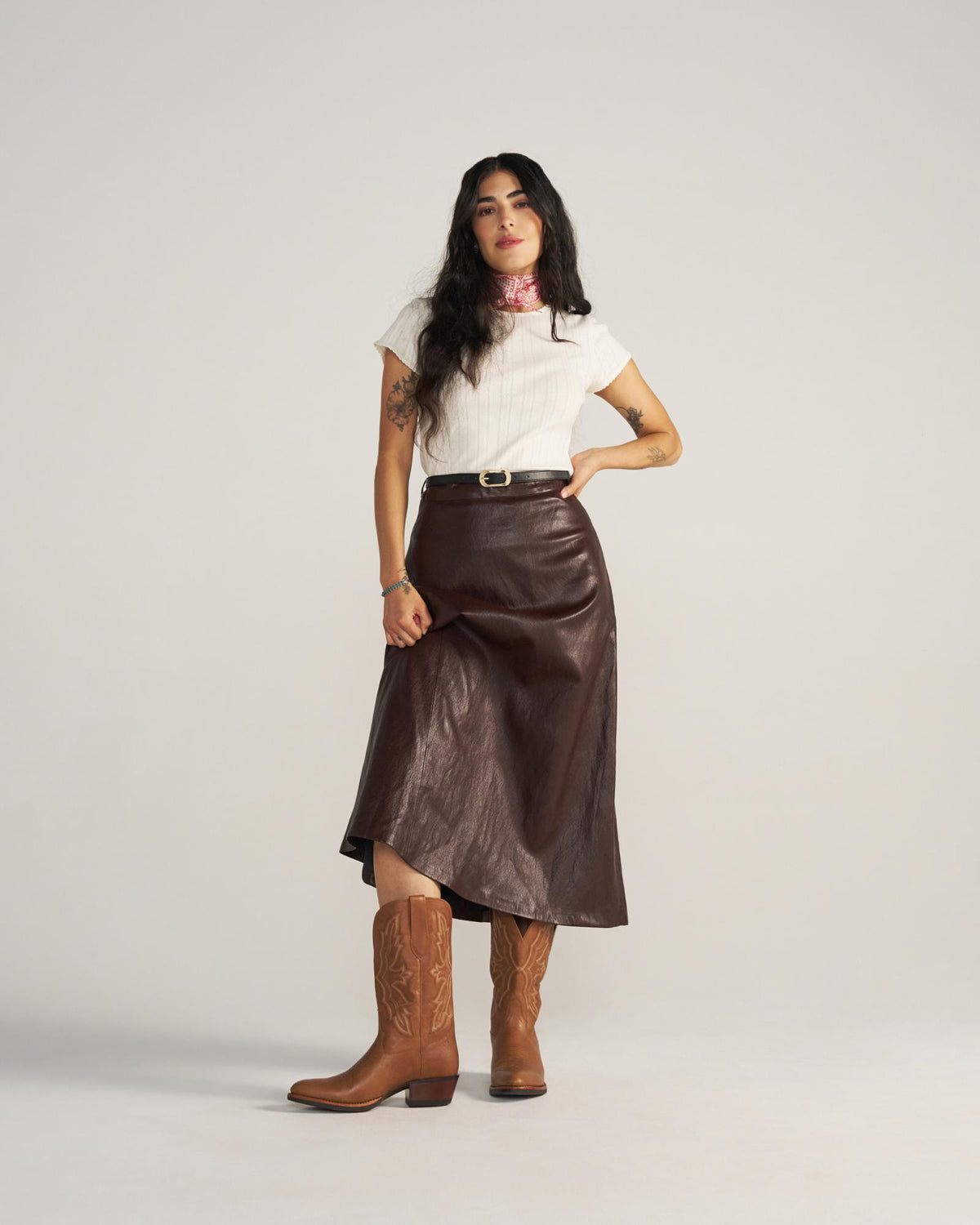 A woman stands against a plain background wearing a white short-sleeve top, brown leather skirt, brown belt, and brown cowboy boots—a chic look inspired by The Britt Goat’s signature latte tones.