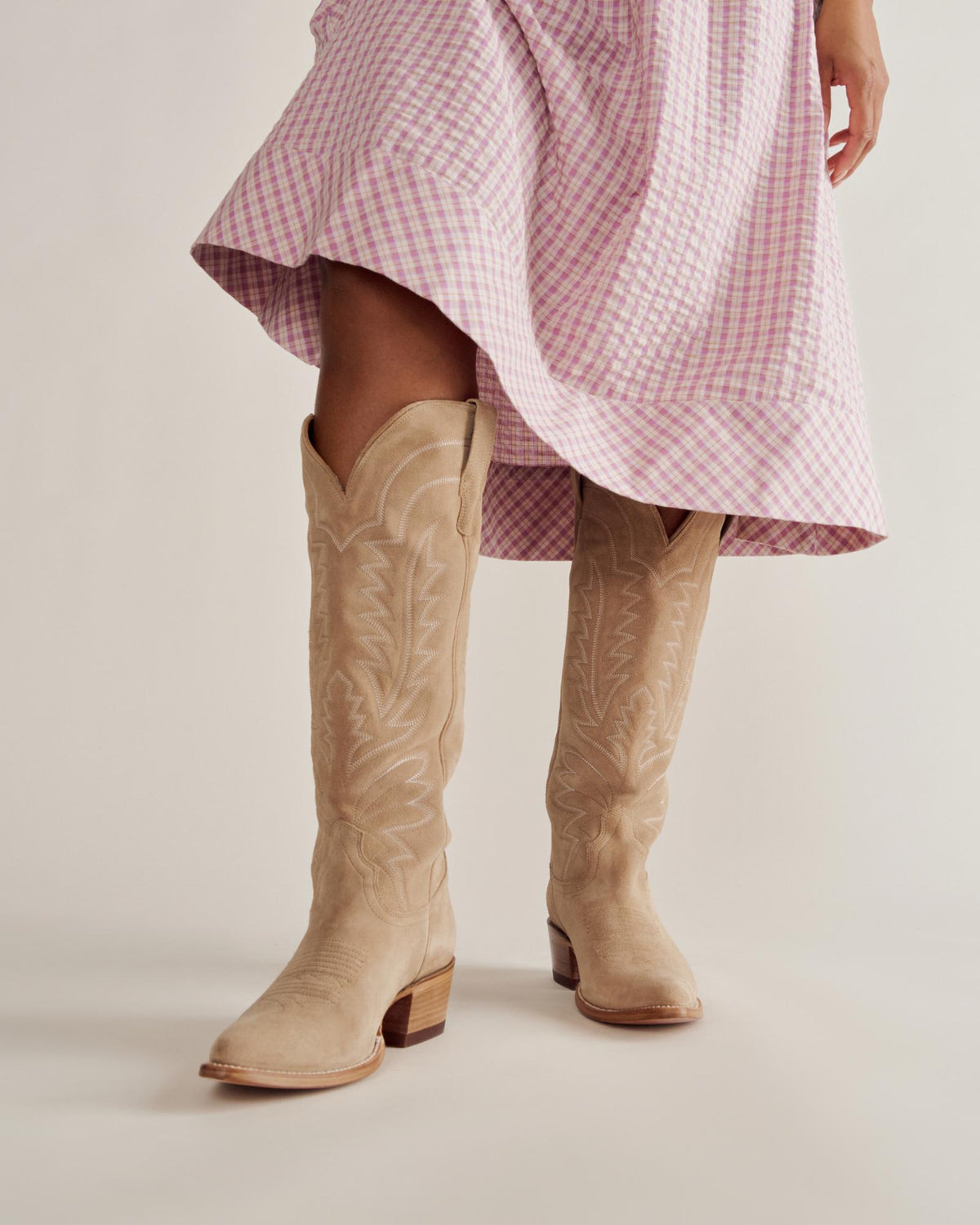 A person wearing a pink checkered dress and beige knee-high cowboy boots stands against a plain background.