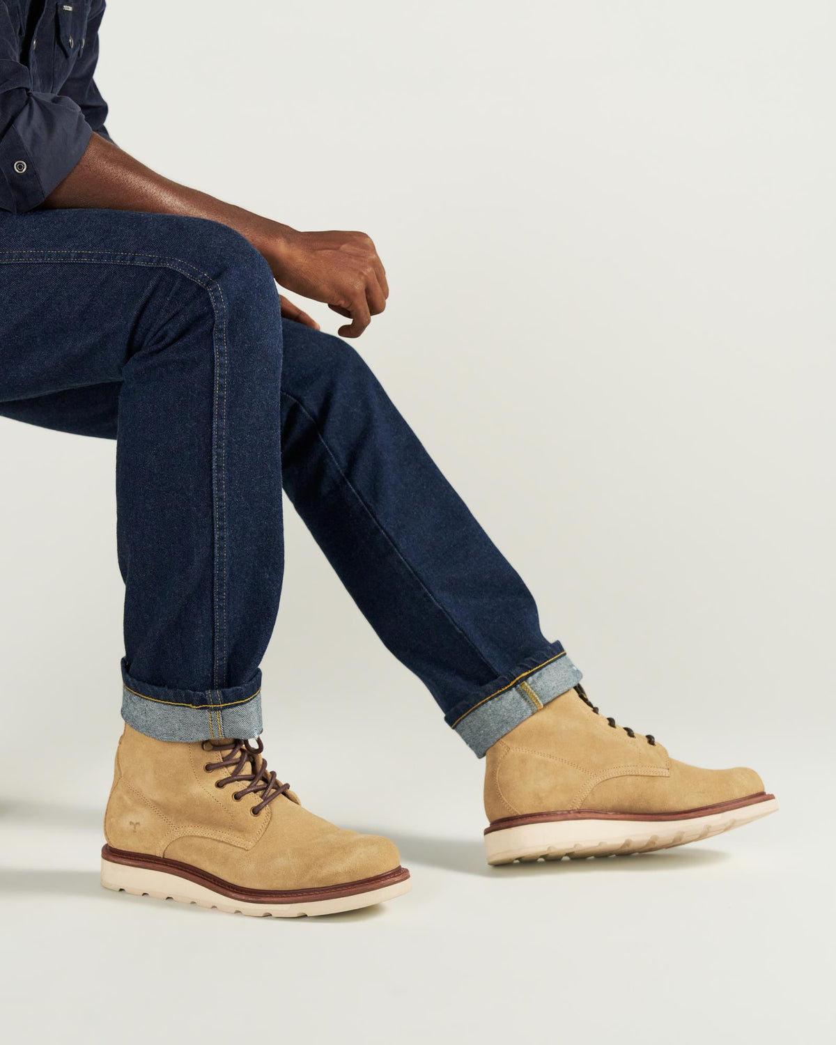 The Knox Lace Up Shearling in Sandstone Roughout. Person sitting on a stool wears dark blue jeans with cuffed hems and tan Sandstone Roughout lace-up boots against a plain, light background.