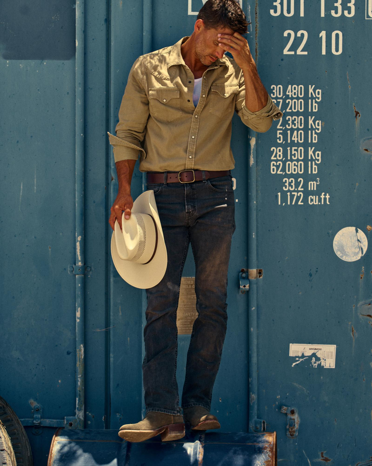 A man in a tan shirt and jeans stands in front of a blue shipping container, holding a white cowboy hat and covering part of his face with his hand.