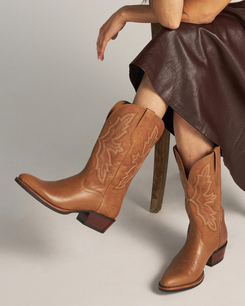 The Britt in Latte Goat. Person sitting in a chair wearing a dark brown skirt and tan cowboy boots with decorative stitching, sipping a creamy Latte Goat coffee.