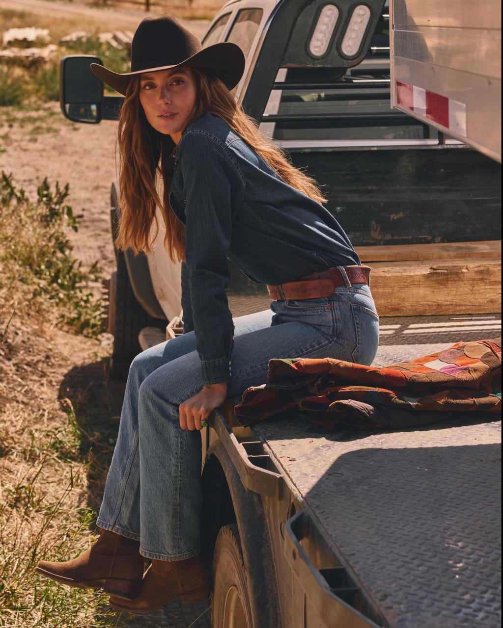 A person wearing a cowboy hat and denim outfit sits on the back of a truck parked on a dirt path.