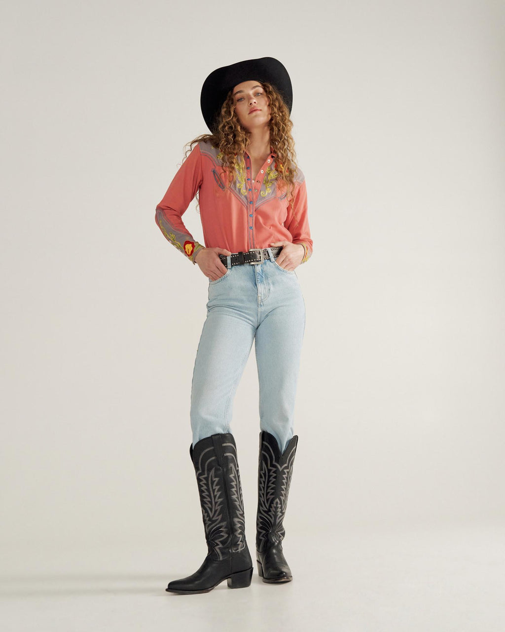 Person wearing a black cowboy hat, embroidered pink shirt, light blue jeans, and black cowboy boots stands against a plain background.