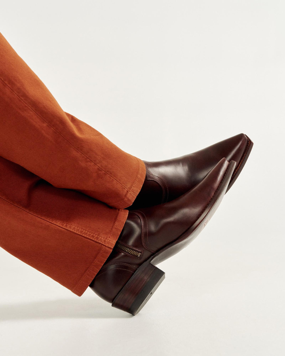Person wearing brown dress shoes and orange pants, with legs crossed against a plain background.