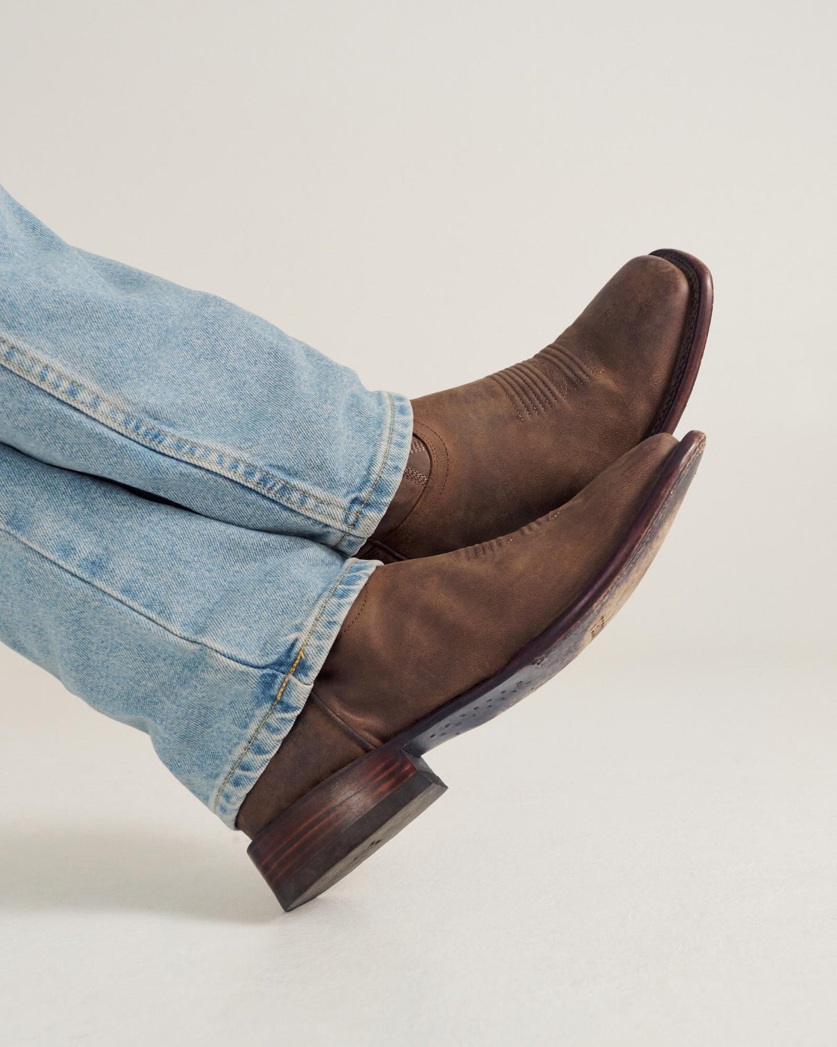 Person wearing light blue jeans and brown leather cowboy boots with legs crossed against a plain background.