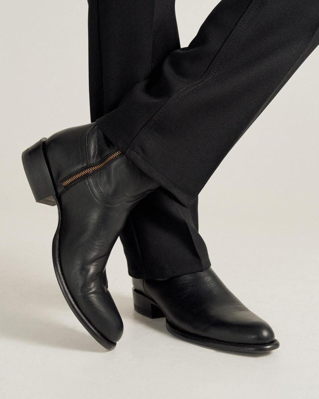 A person wearing black dress pants and black leather boots with a side zipper, standing with one leg crossed over the other.