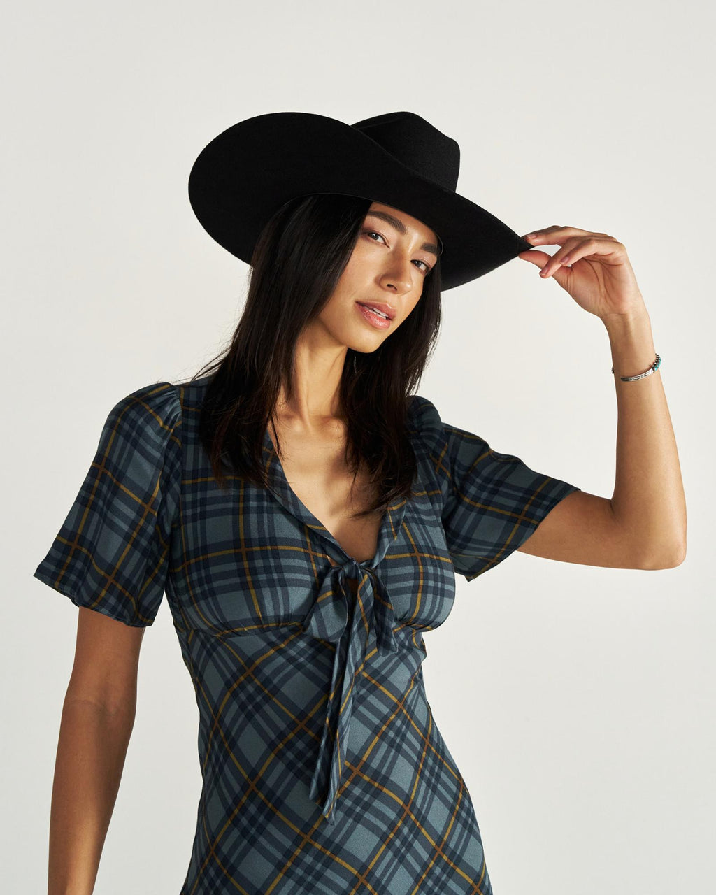 Woman wearing a black cowboy hat and a plaid dress stands against a plain background, holding the brim of her hat with one hand.