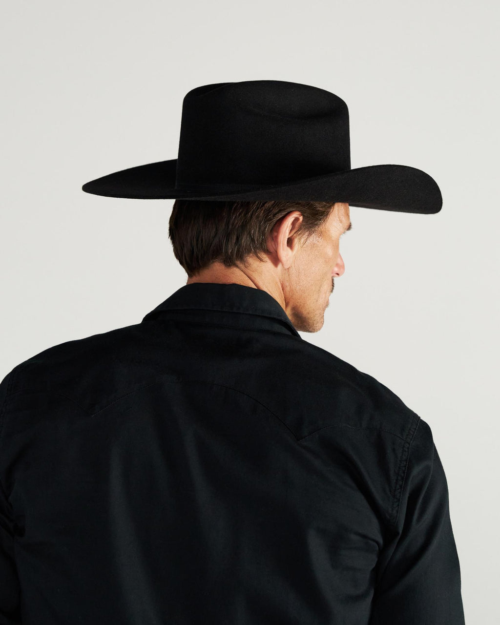 Man wearing a black wide-brimmed hat and black shirt, shown from behind against a plain, light background.