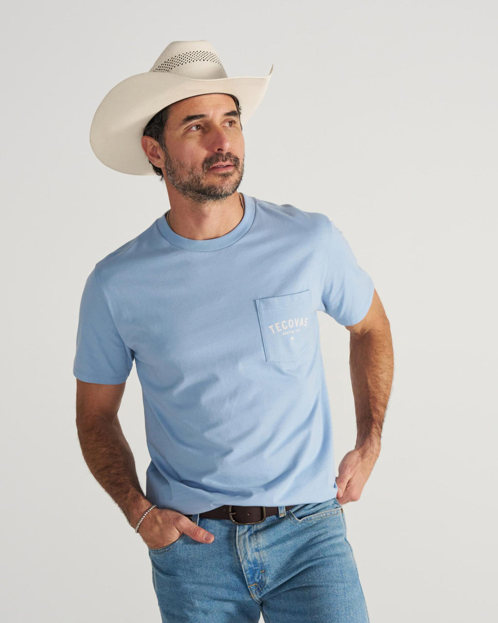 A man in a light blue T-shirt, jeans, and a white cowboy hat stands with one hand on his hip, looking to the side against a plain background.