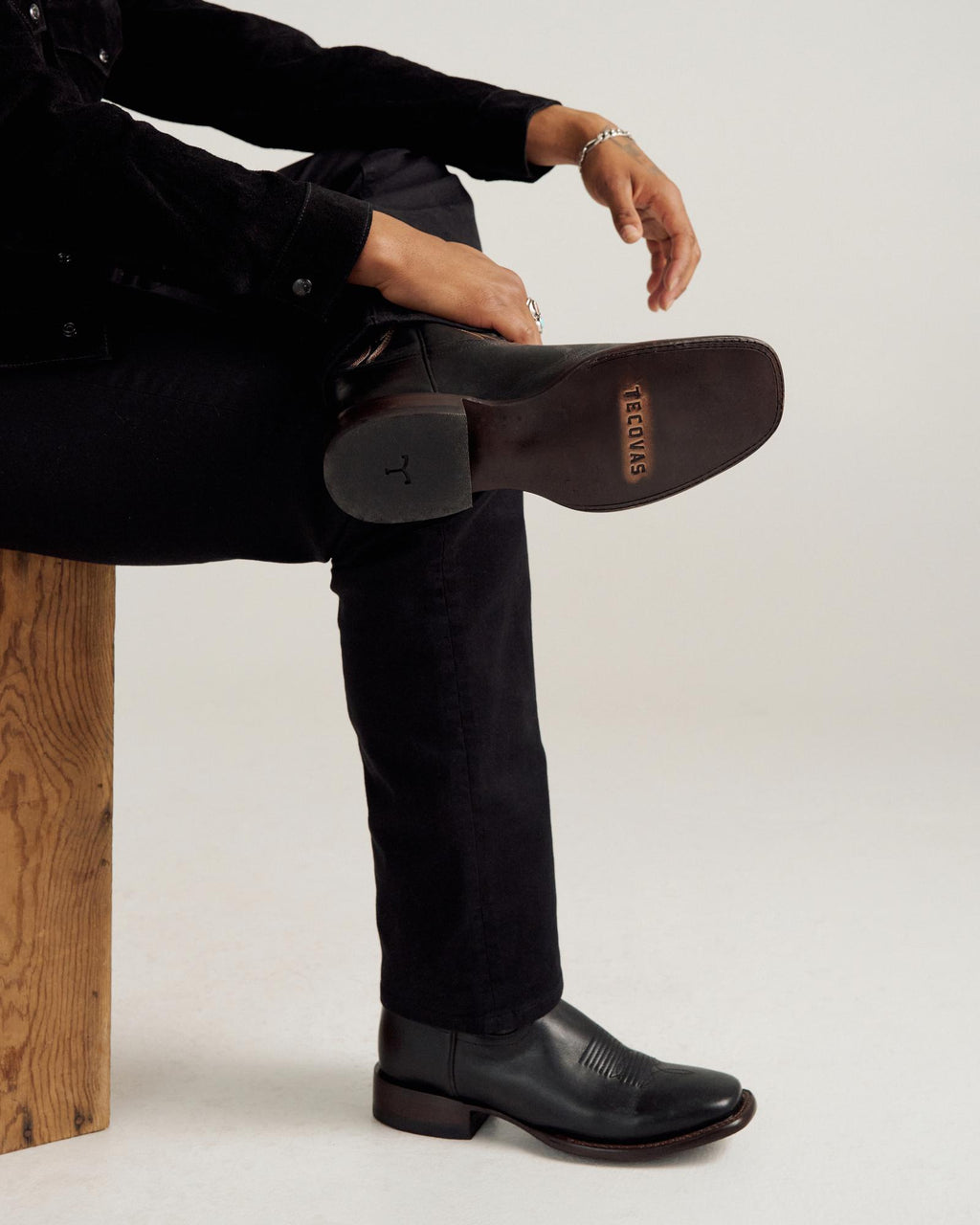 A person in black pants and boots sits on a wooden bench with one leg crossed, showing the sole of the boot stamped with the word “TEXAS.”.